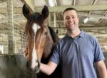Smiling man in a blue polo beside a brown horse in a barn, hand resting on the horse's neck.