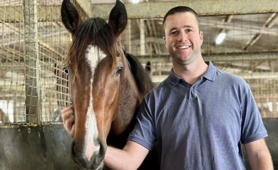 Smiling man in a blue polo beside a brown horse in a barn, hand resting on the horse's neck.