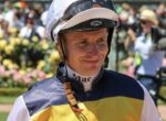Smiling jockey in yellow and navy silks with helmet on, at a sunny racecourse with blurred spectators behind him.