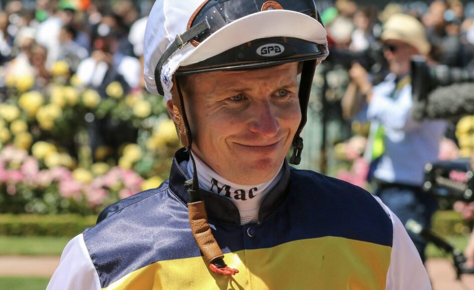 Smiling jockey in yellow and navy silks with helmet on, at a sunny racecourse with blurred spectators behind him.