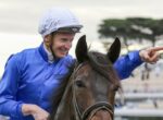 Jockey in blue silks and white helmet riding a brown horse, smiling as he points forward off-camera at a racecourse.