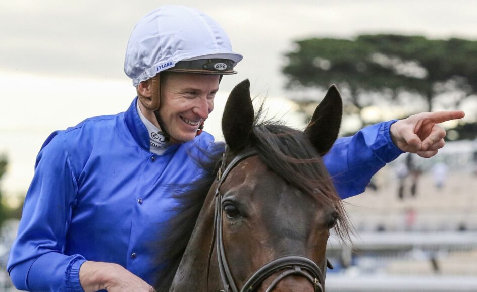 Jockey in blue silks and white helmet riding a brown horse, smiling as he points forward off-camera at a racecourse.