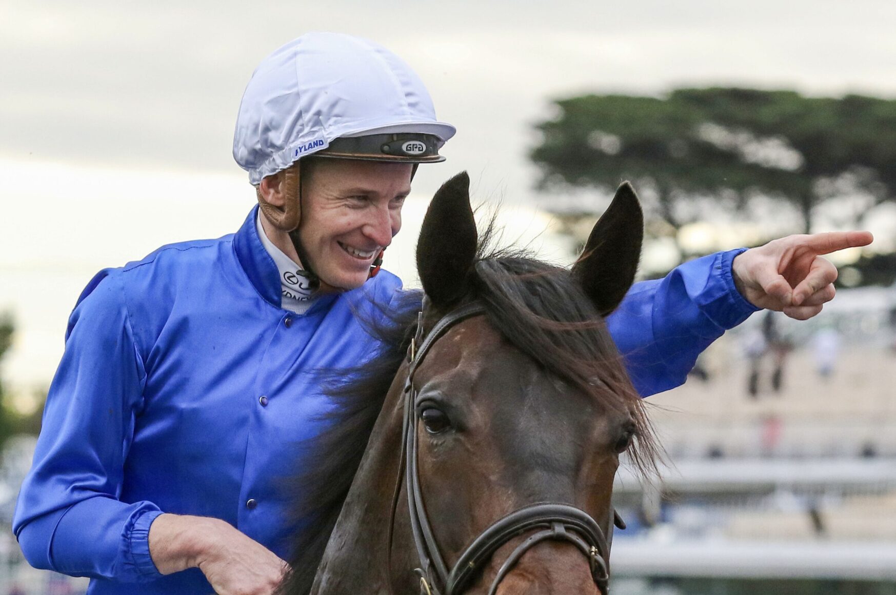 Jockey in blue silks and white helmet riding a brown horse, smiling as he points forward off-camera at a racecourse.