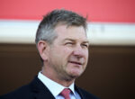 Head-and-shoulders portrait of a middle-aged man in a dark suit and red tie, outdoors against a red backdrop.