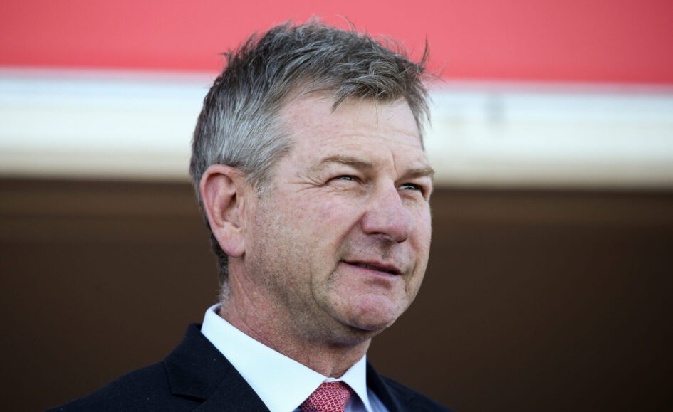 Head-and-shoulders portrait of a middle-aged man in a dark suit and red tie, outdoors against a red backdrop.