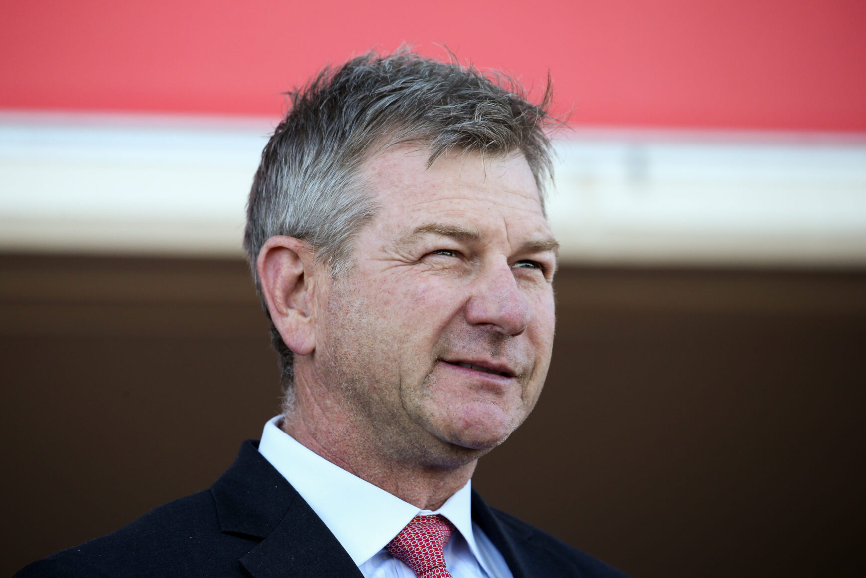 Head-and-shoulders portrait of a middle-aged man in a dark suit and red tie, outdoors against a red backdrop.