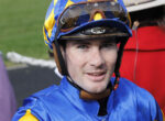 Close-up of a jockey in blue and yellow helmet gear, smiling at the camera with goggles on top of the helmet.