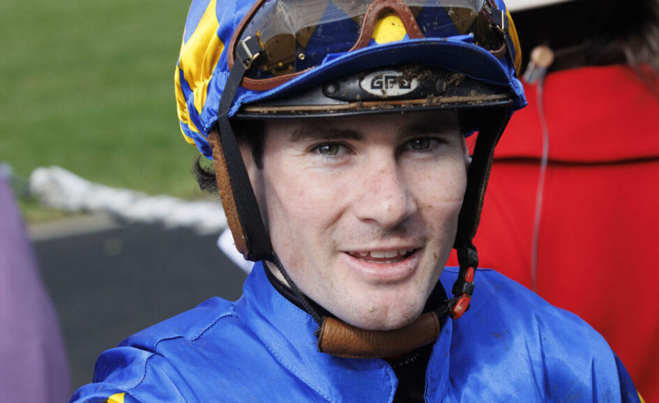 Close-up of a jockey in blue and yellow helmet gear, smiling at the camera with goggles on top of the helmet.