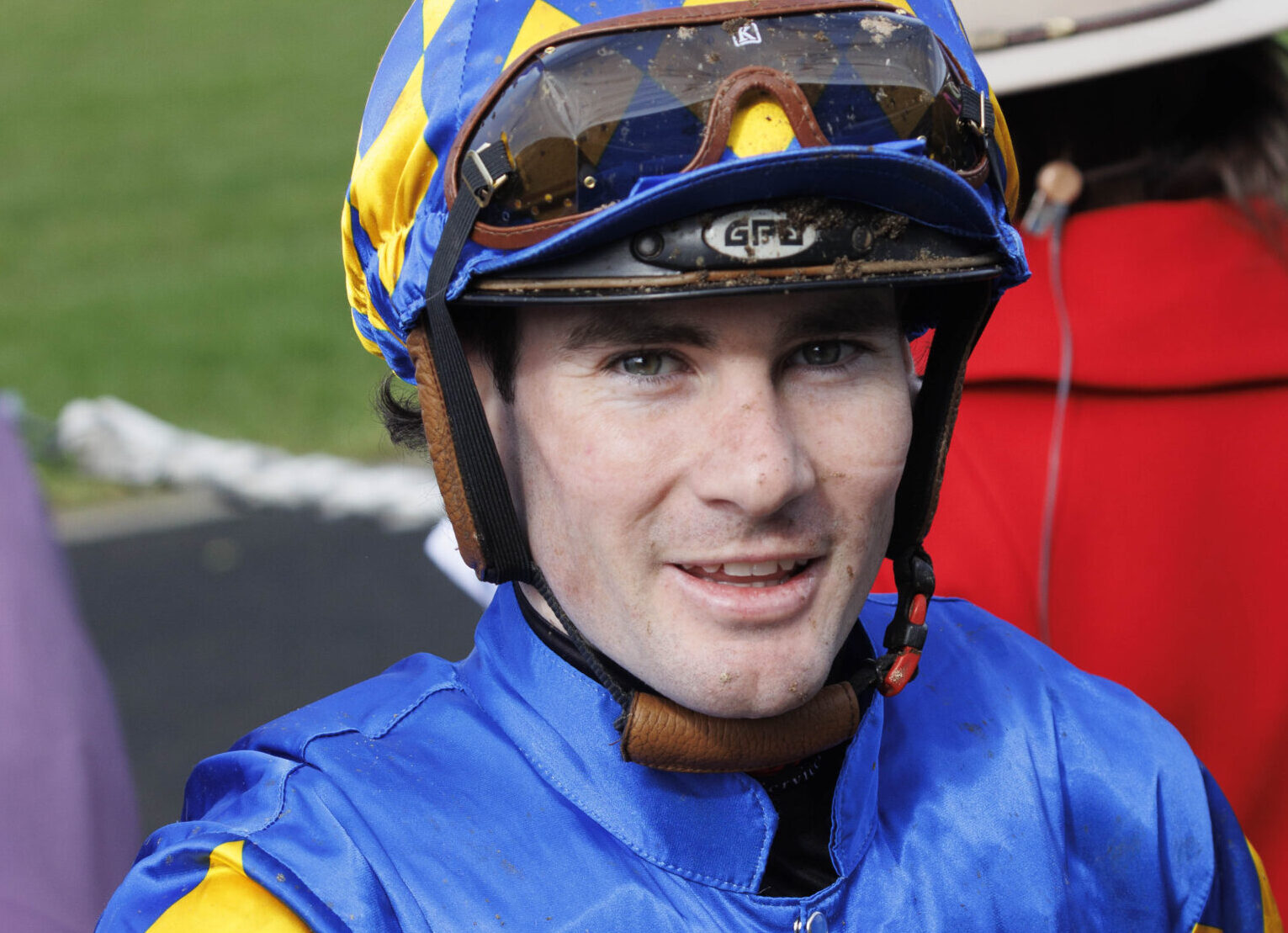 Close-up of a jockey in blue and yellow helmet gear, smiling at the camera with goggles on top of the helmet.