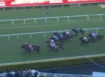 Group of jockeys and horses racing on a green turf track, colorful silks flashing as they round the bend near the railings, with spectators in the foreground.