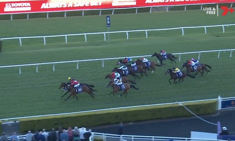Group of jockeys and horses racing on a green turf track, colorful silks flashing as they round the bend near the railings, with spectators in the foreground.