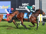 Two jockeys and their horses race side by side on a grassy track at Caulfield Racecourse, with signage in the background.
