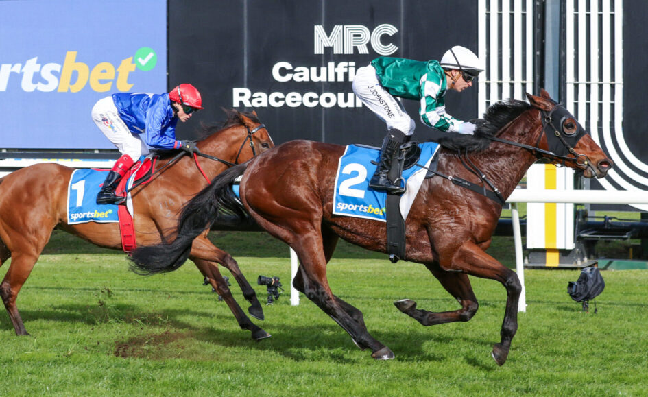 Two jockeys and their horses race side by side on a grassy track at Caulfield Racecourse, with signage in the background.