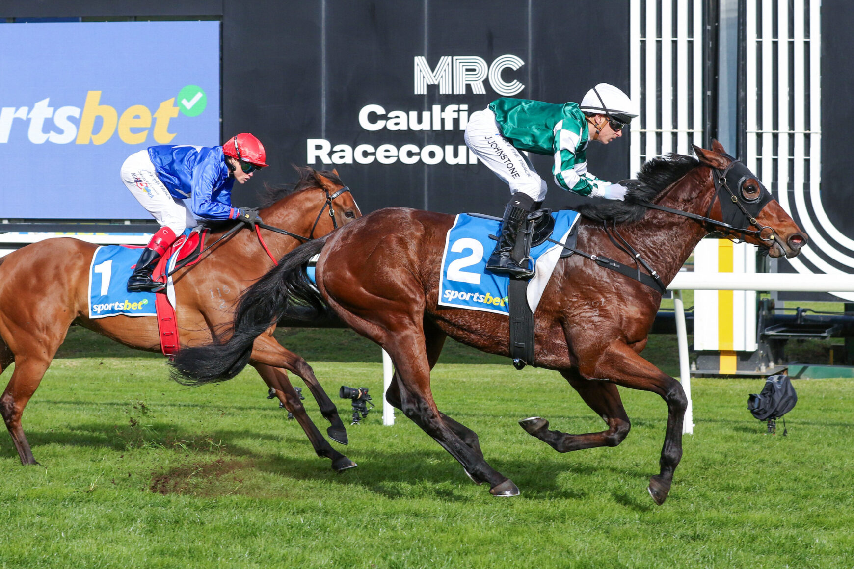Two jockeys and their horses race side by side on a grassy track at Caulfield Racecourse, with signage in the background.