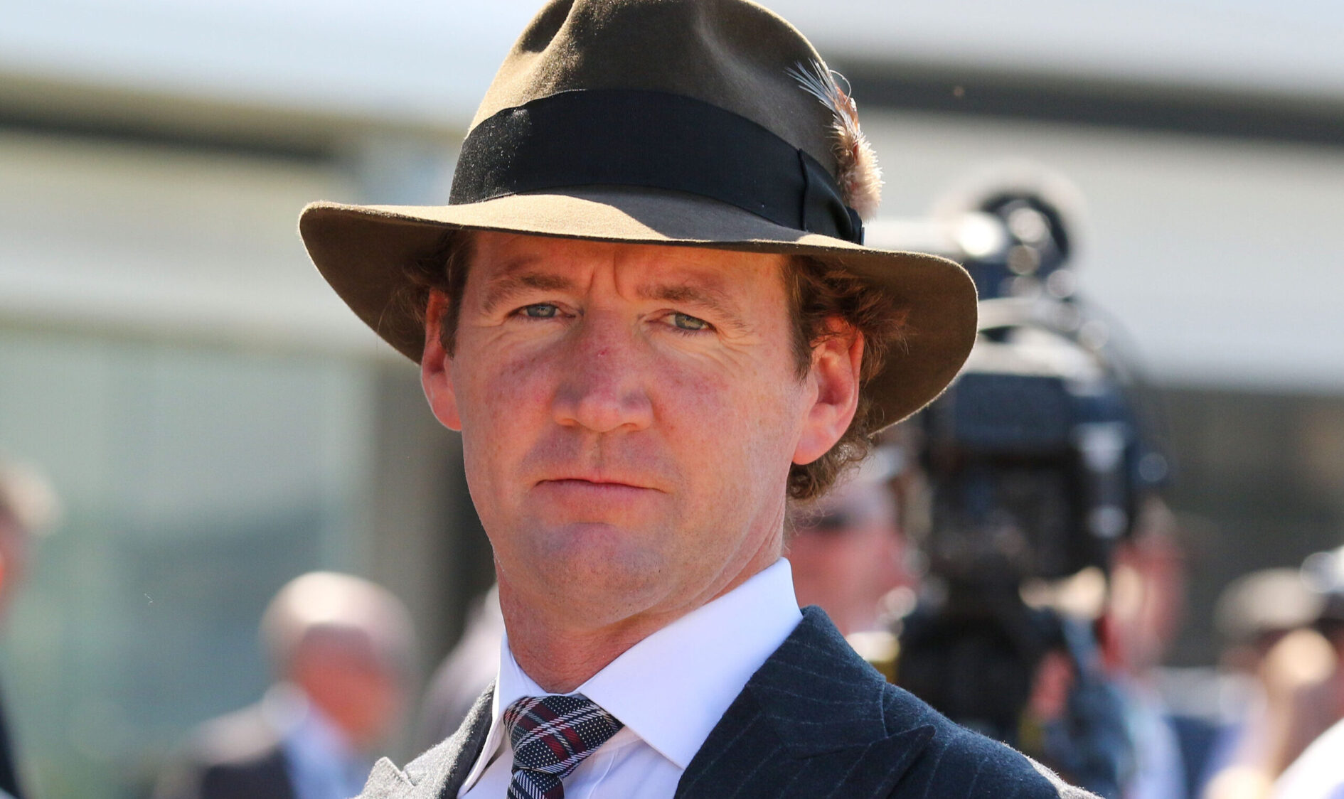 Close-up of a man in a pinstripe suit and brown fedora at an outdoor event.