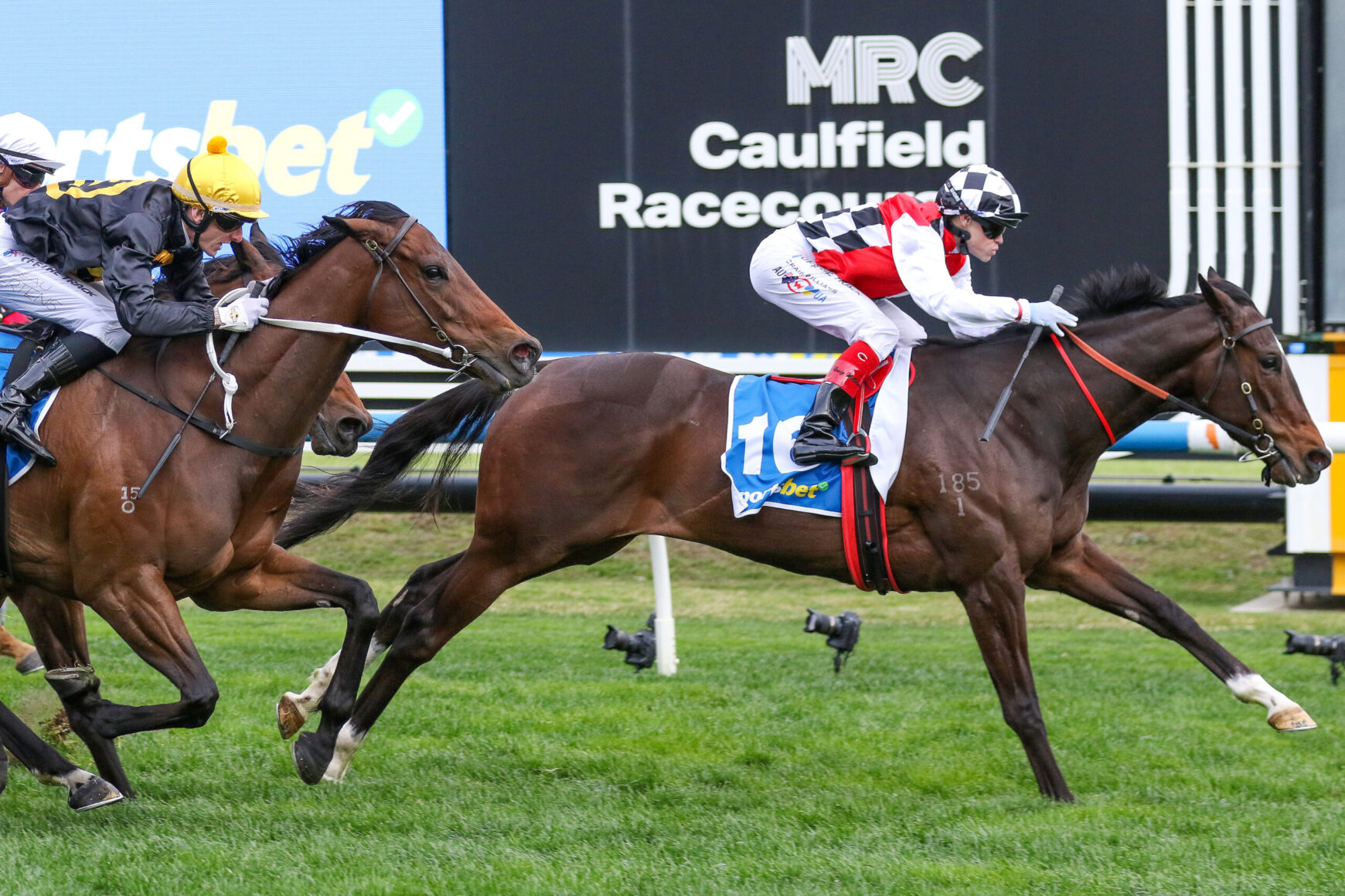 Two jockeys ride racing horses at Caulfield Racecourse during a close finish on a sunny day.