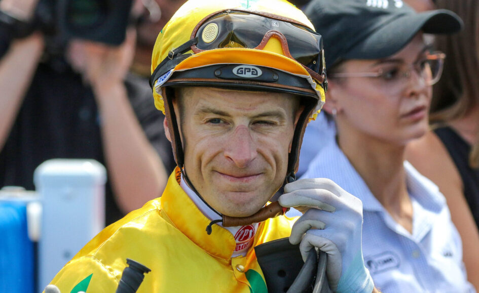 Race car driver in a bright yellow suit and helmet with goggles, outdoors among spectators.