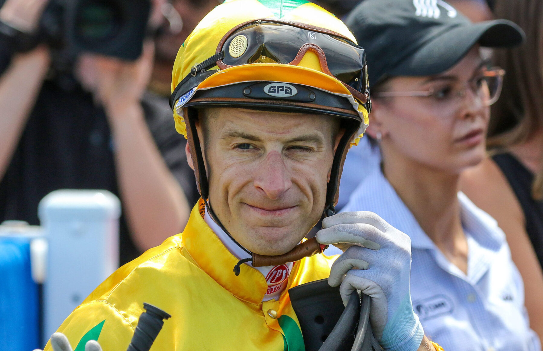 Race car driver in a bright yellow suit and helmet with goggles, outdoors among spectators.
