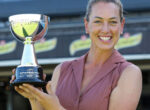 Smiling woman in a sleeveless blouse holds a large silver trophy at a sports stadium. It’s an awards moment for a winner.
