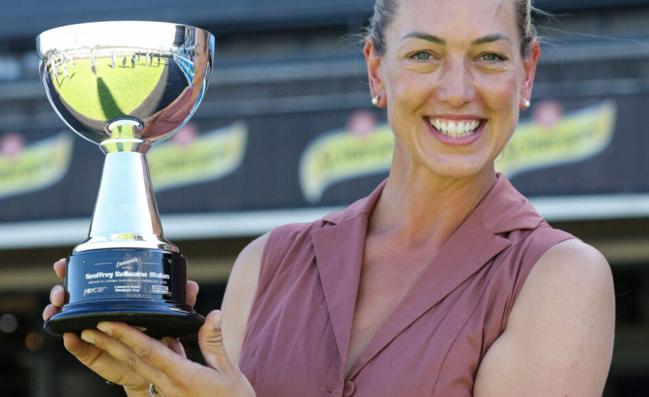 Smiling woman in a sleeveless blouse holds a large silver trophy at a sports stadium. It’s an awards moment for a winner.