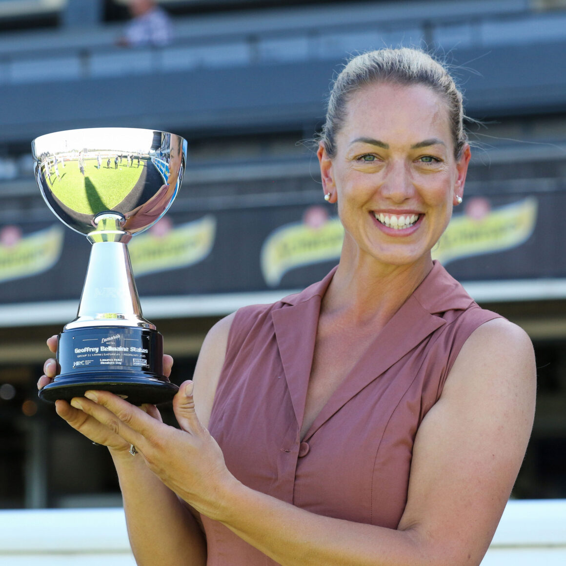 Smiling woman in a sleeveless blouse holds a large silver trophy at a sports stadium. It’s an awards moment for a winner.