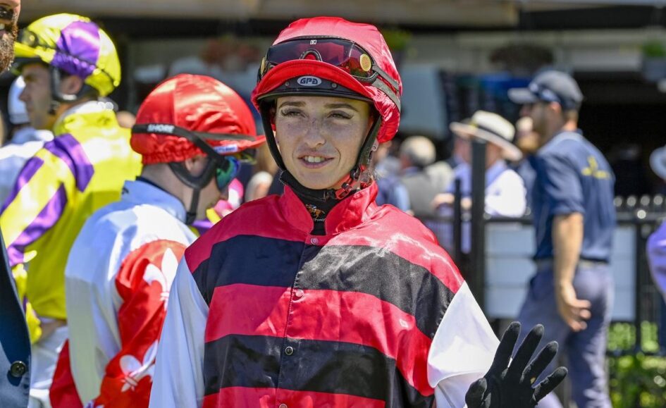 Woman jockey in red-and-black striped silks with a red helmet waves, surrounded by other riders in colorful gear at a paddock.
