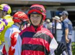 Jockey in red-and-black striped silks waves hello at a busy paddock, wearing a red helmet and black gloves.