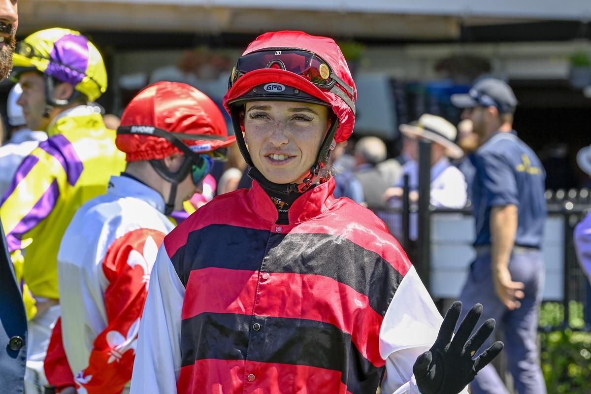Jockey in red-and-black striped silks waves hello at a busy paddock, wearing a red helmet and black gloves.