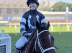 Smiling jockey in blue-and-black racing silks rides a dark horse on a racecourse side view.