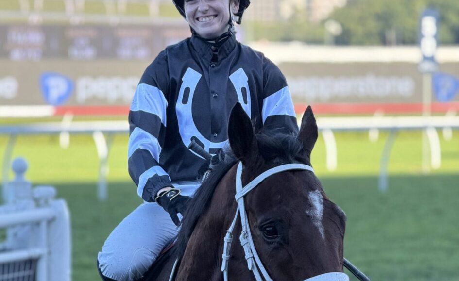 Smiling jockey in blue-and-black racing silks rides a dark horse on a racecourse side view.