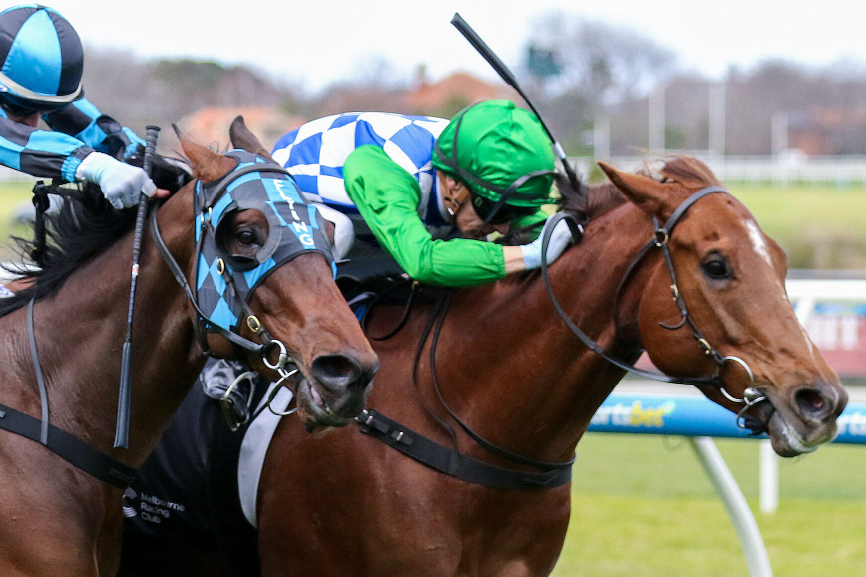 Two jockeys ride competing horses neck-and-neck in a race, one in green silks and the other in blue checkered silks, on a grassy track.