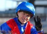 Smiling jockey wearing a blue helmet and red-blue racing silks, waving with a gloved hand.