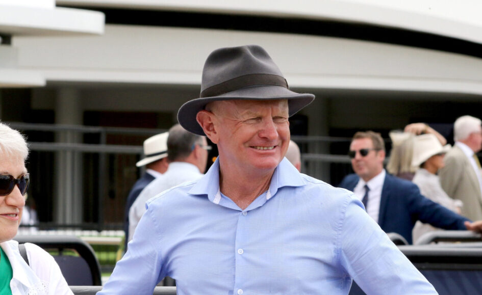 Smiling man wearing a gray fedora and light blue shirt at an outdoor event, with other guests in the background.
