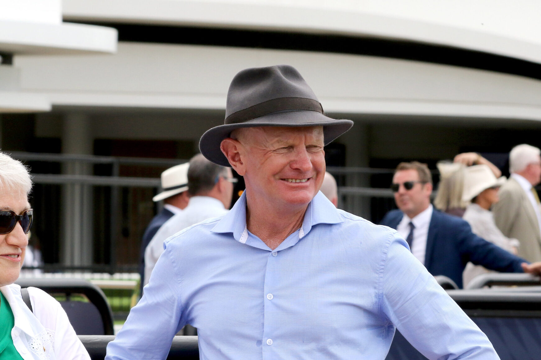 Smiling man wearing a gray fedora and light blue shirt at an outdoor event, with other guests in the background.