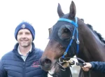 Man in a blue quilted jacket and knit hat stands beside a brown horse wearing a blue bridle, both looking toward the camera.