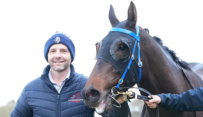 Man in a blue quilted jacket and knit hat stands beside a brown horse wearing a blue bridle, both looking toward the camera.