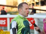 Jockey in green racing silks being interviewed by a reporter with a red microphone at a racecourse surface behind them.