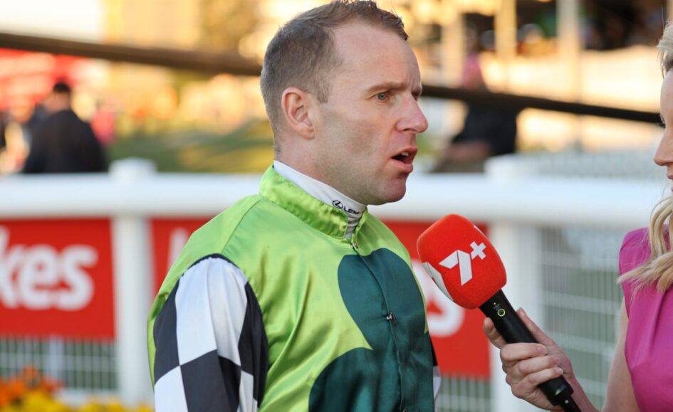 Jockey in green racing silks being interviewed by a reporter with a red microphone at a racecourse surface behind them.