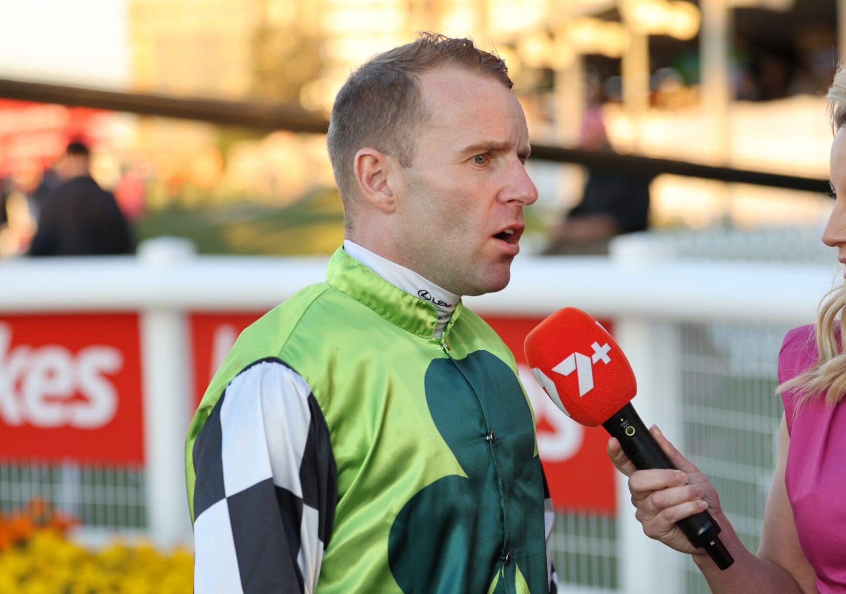 Jockey in green racing silks being interviewed by a reporter with a red microphone at a racecourse surface behind them.