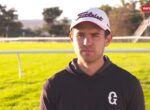 Male athlete wearing a white Titleist cap and black hoodie on a sunny racecourse with a green infield.