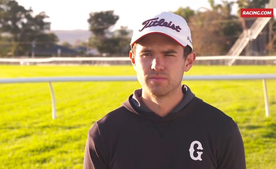 Male athlete wearing a white Titleist cap and black hoodie on a sunny racecourse with a green infield.