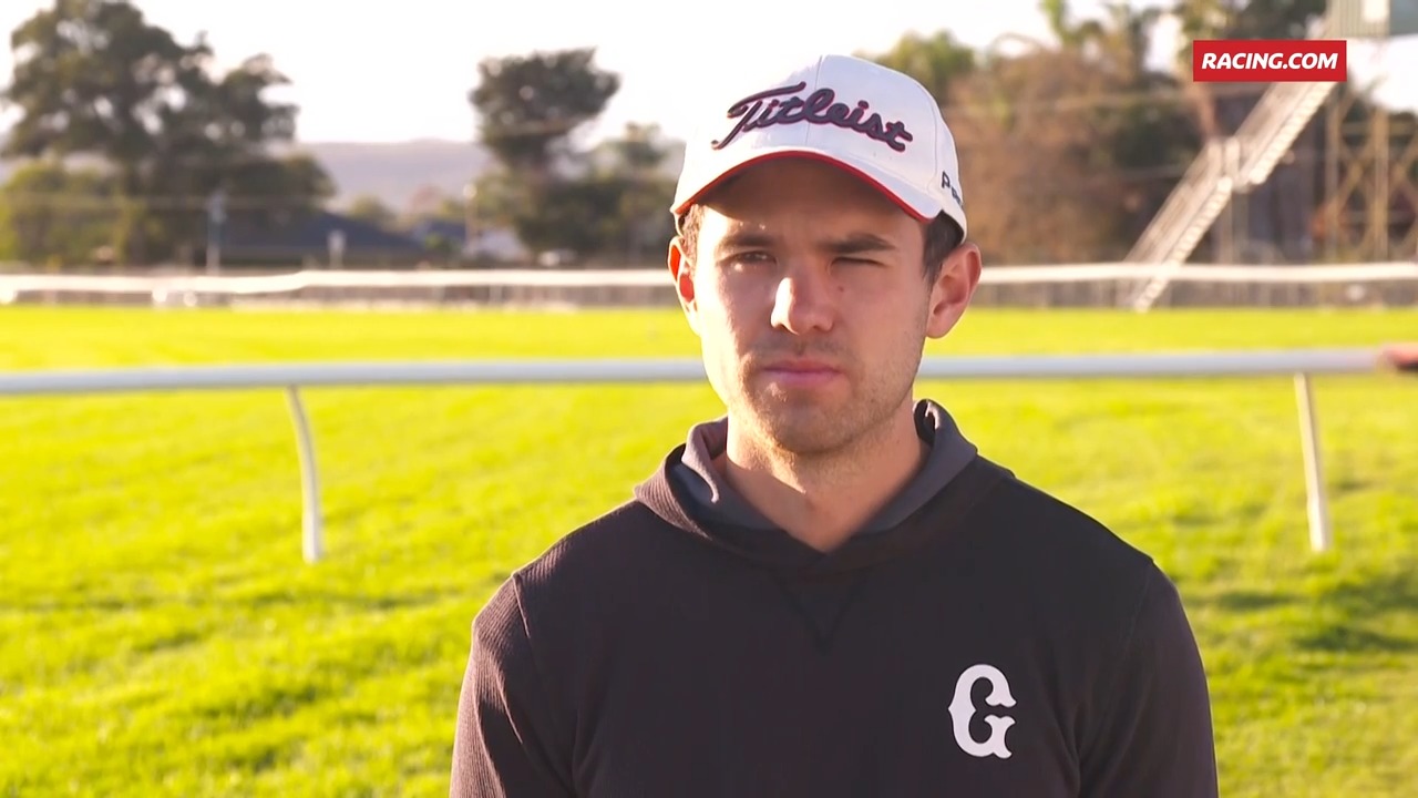 Male athlete wearing a white Titleist cap and black hoodie on a sunny racecourse with a green infield.