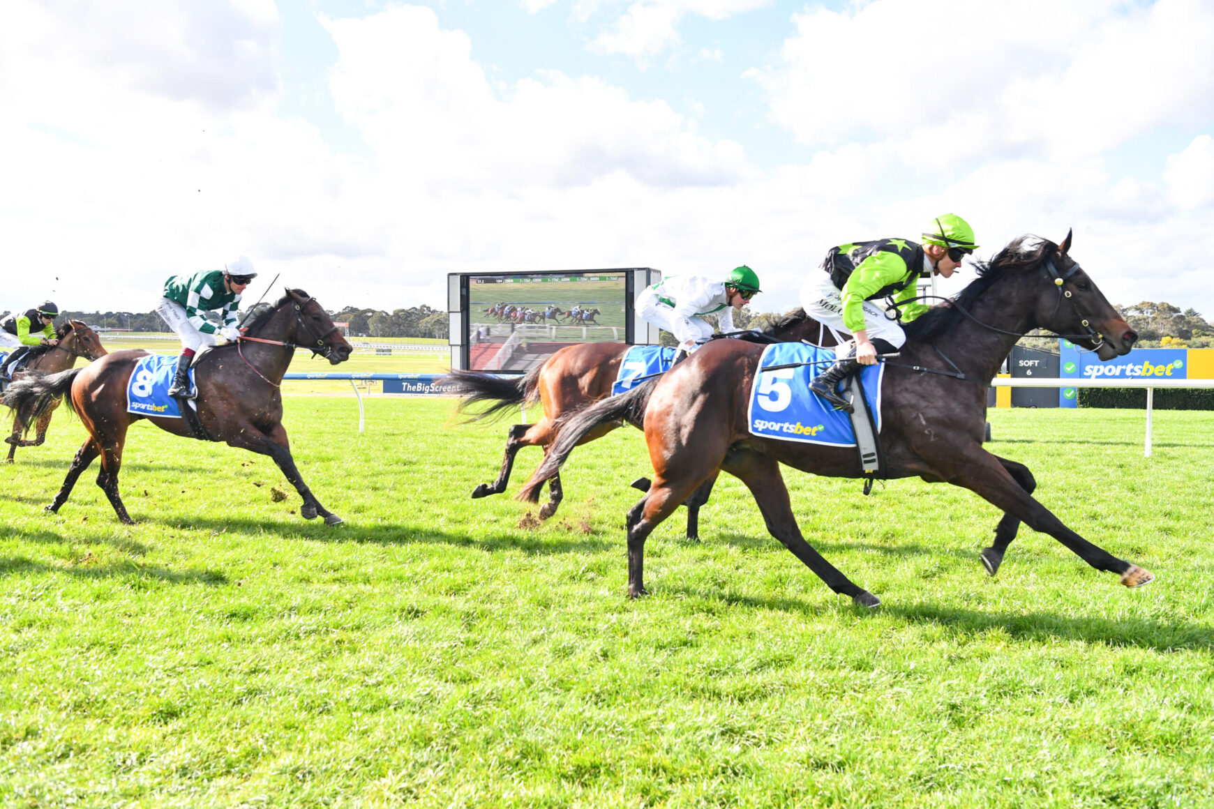 Jockeys and horses race on a sunny track, closest to camera is a dark horse with a neon green cap, blue saddlecloths visible, crowd and scoreboard in background