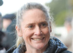 Close-up portrait of a smiling woman with gray hair tied back, wearing a dark jacket outdoors.