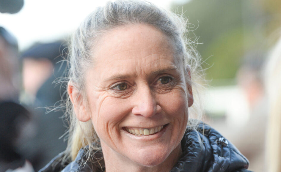 Close-up portrait of a smiling woman with gray hair tied back, wearing a dark jacket outdoors.