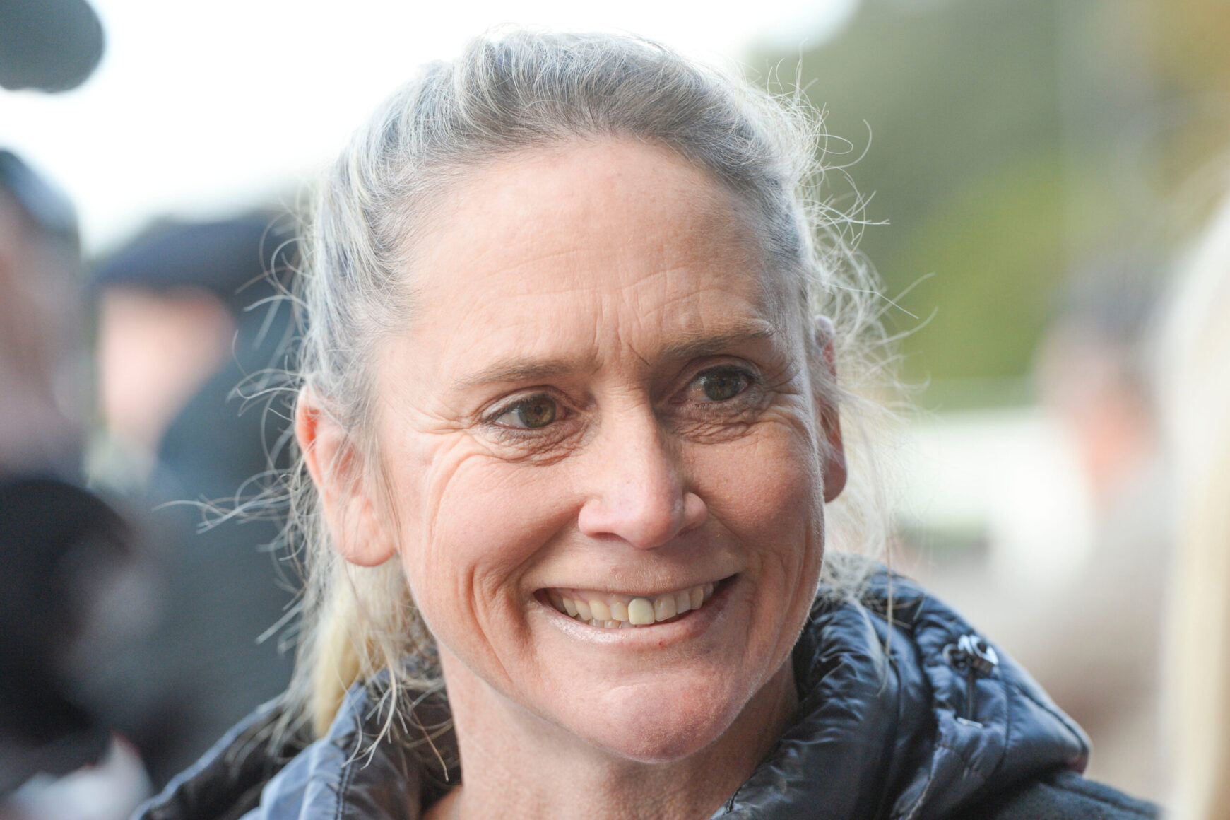 Close-up portrait of a smiling woman with gray hair tied back, wearing a dark jacket outdoors.