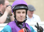 Portrait of a jockey in a purple helmet and jacket, smiling at the camera and making a peace sign.