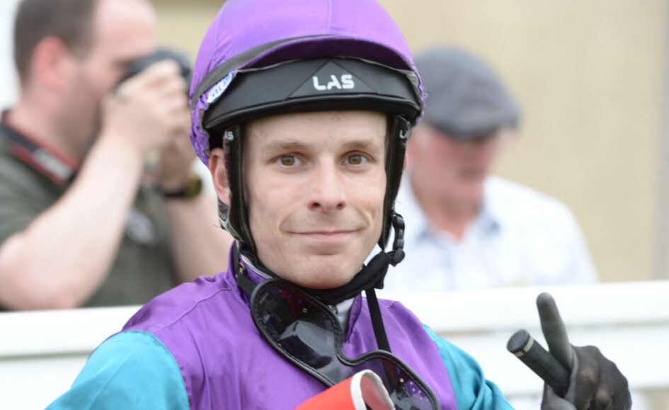 Portrait of a jockey in a purple helmet and jacket, smiling at the camera and making a peace sign.