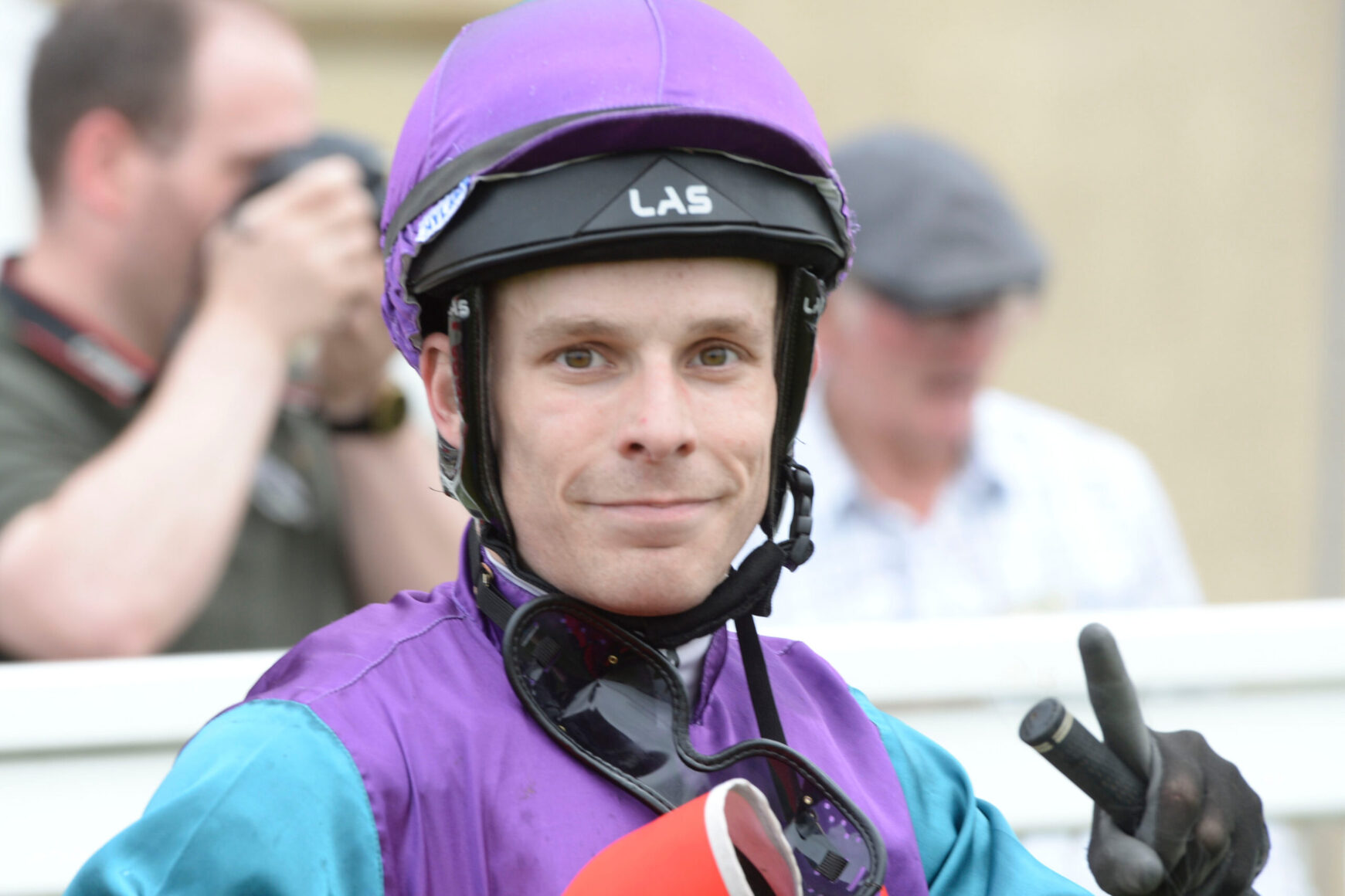 Portrait of a jockey in a purple helmet and jacket, smiling at the camera and making a peace sign.