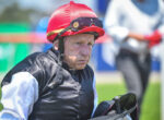 Older jockey wearing a red helmet and black riding vest, holding a racing saddle, outdoors on a sunny day